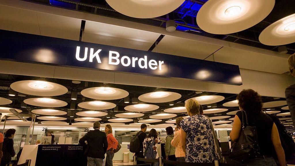 UK airport passport control. There is a blue overhead sign with UK border written on it in white. People are queuing to get their passports checked.