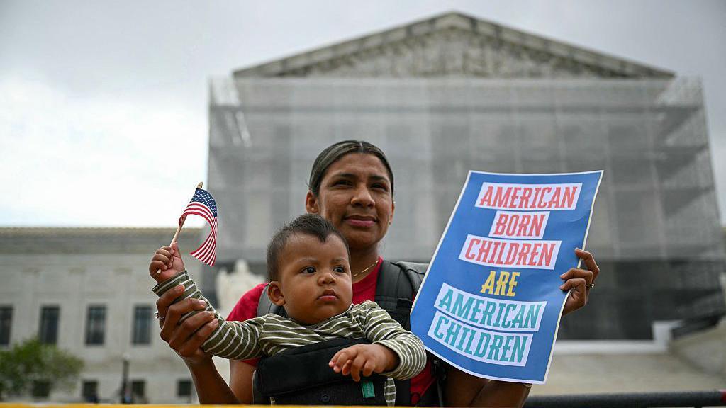 A mother with her infant son hold a US flag and sign saying "American born children are American children" outside the US Supreme Court