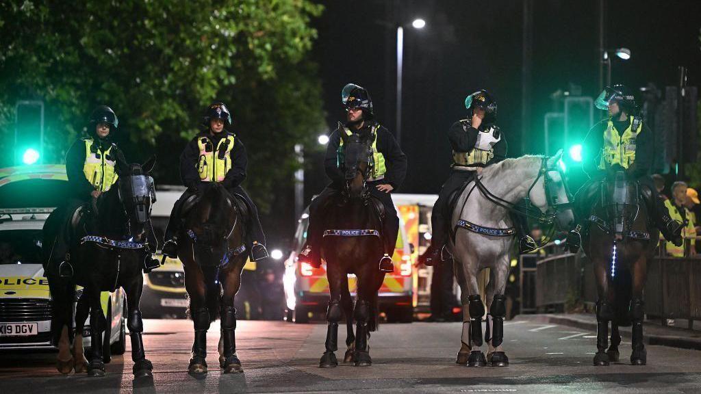 A line of police officers on horses at night. They are all wearing hi-vis vests and helmets. In the background you can see several police vans parked in the middle of the road.