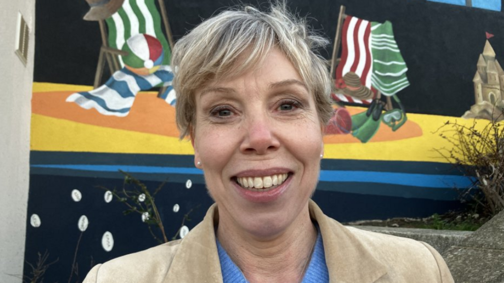 A woman with short blonde hair smiles as she stands in front of a brightly coloured mural depicting striped deckchairs, striped towels and a sandcastle on a beach. She is wearing pearl stud earrings, a beige jacket and a blue top.