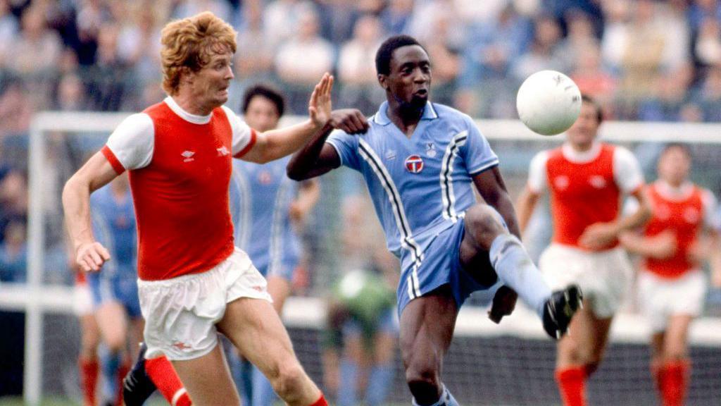 Two men are playing football and wearing football kits while standing on a football pitch.