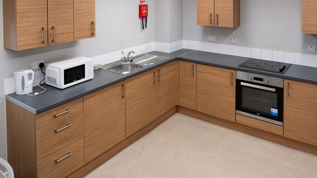 A kitchen with a pale floor, wooden units and a black worktop. There is an oven, a kettle and a microwave and three wall-mounted units