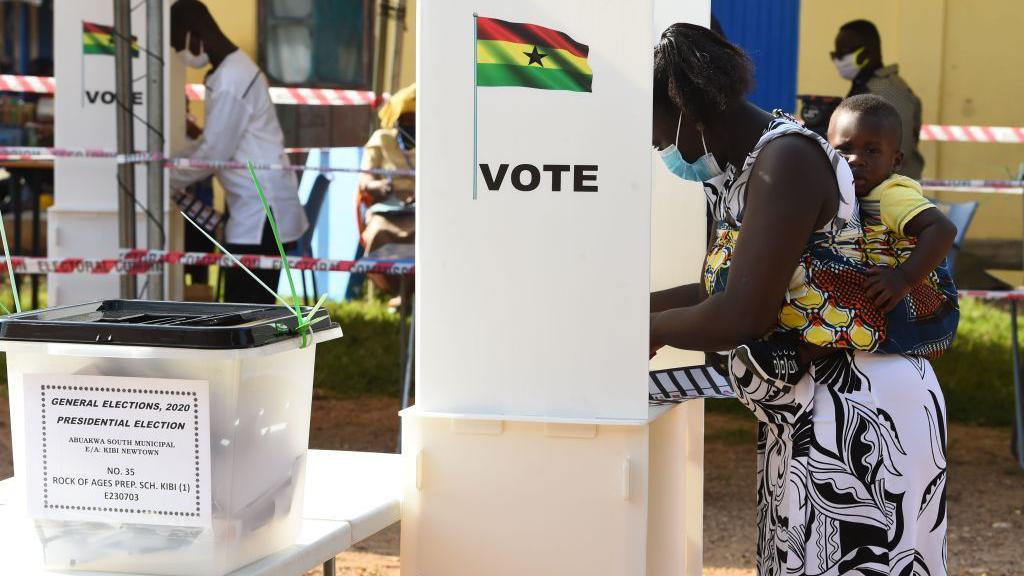 A woman with a baby tied to her back stands at an outdoor polling booth to mark her ballot paper.