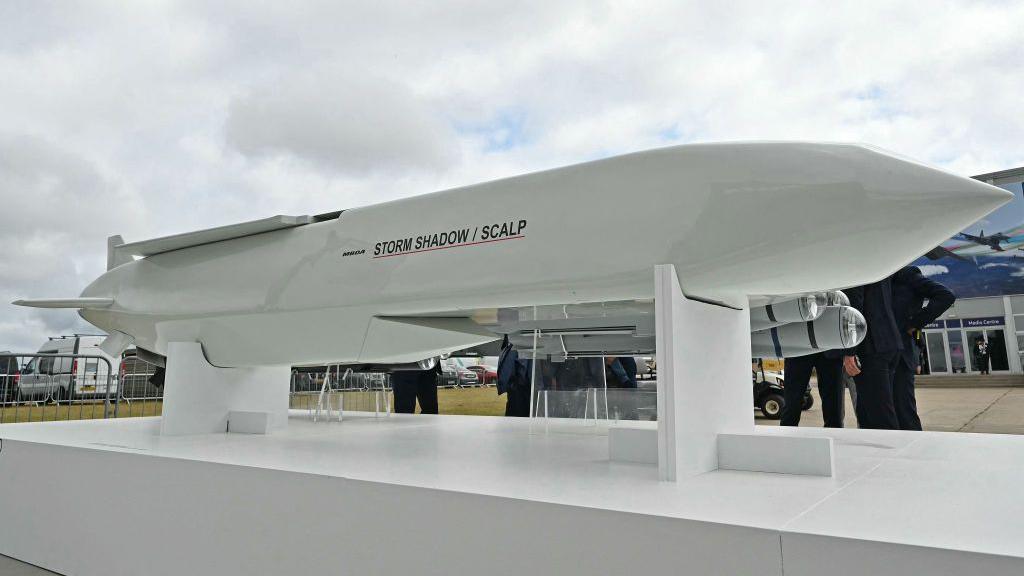 A long white missile on a stand with people stood around it. It has the words storm shadow/scalp on it and has wings at the back and a pointy nose