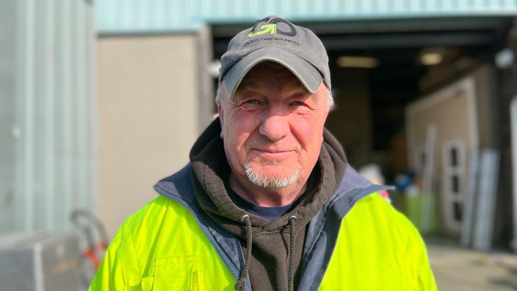 A smiling man with a grey, short beard, wearing a grey cap. He is wearing a black hoodie underneath a yellow fluorescent jacket. 