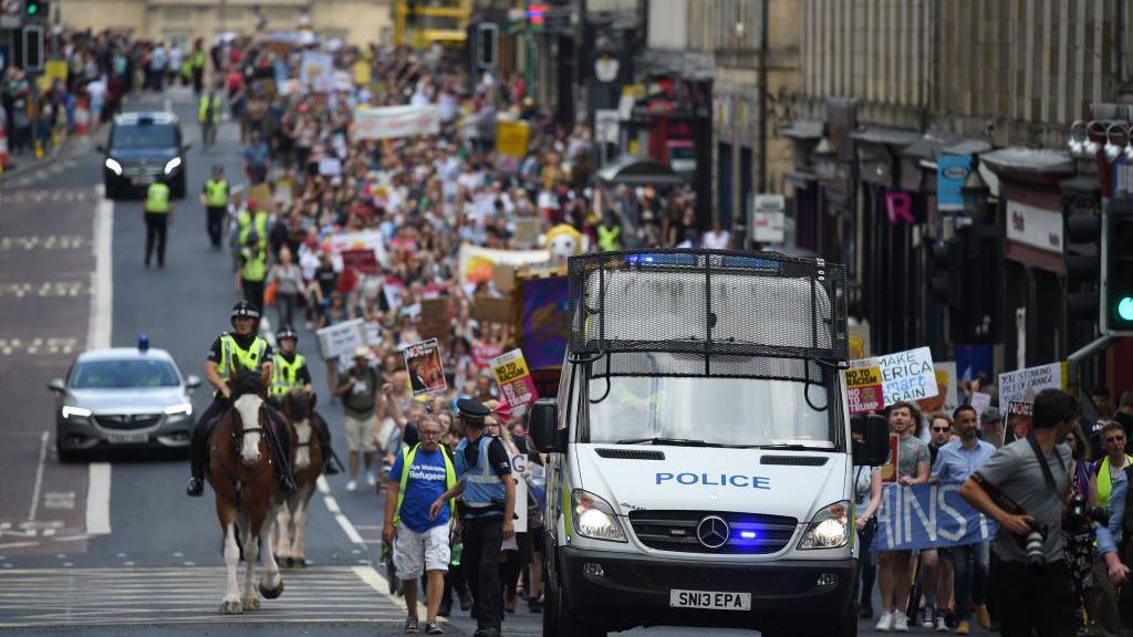 A Police Scotland van followed by a long protest of hundreds of people in a row. They are carrying anti-Trump banners. They are accompanied by mounted police officers.