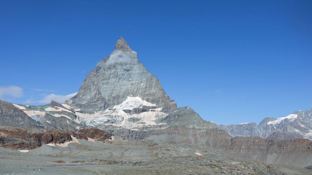 The Matterhorn mountain towering above surrounding ranges, with a tiny cloud near its peak, some snow around the base and a vibrant blue sky.