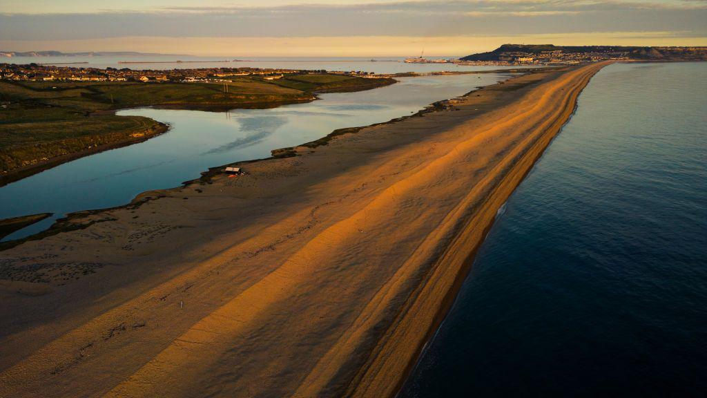 Aerial photo of a battier beach at sunset. There is sea behind and in front of it, with the island of Portland in the background.