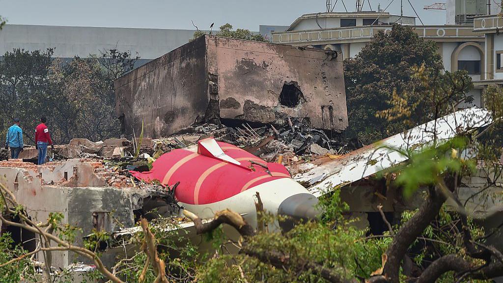 Two investigative officials stand at the site of Air India Boeing 787 crash site. They stand with their backs to the camera, next to the remnants of the plane amid foliage.