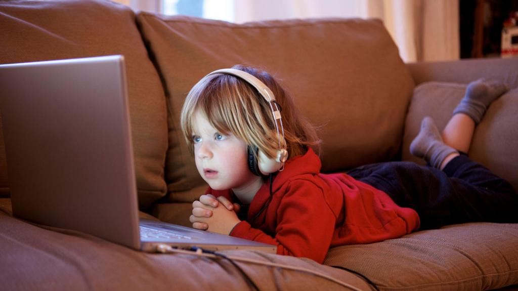 A young child of about five lies on their stomach on a sofa, looking into the glow of a computer screen. they wear large over-ear headphones.
