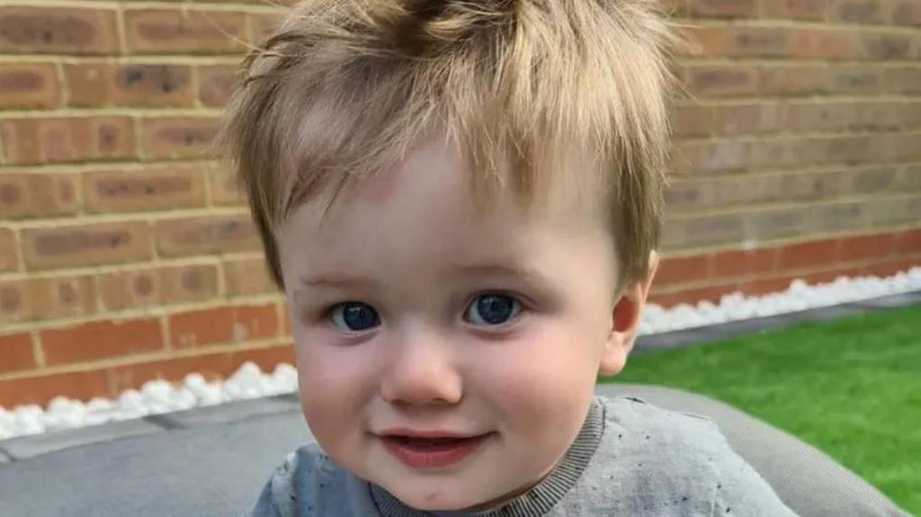 Close up shot of nine-month-old Oliver Steeper outside in front of a wall. He is looking at the camera and has a grey top on and blond hair. 