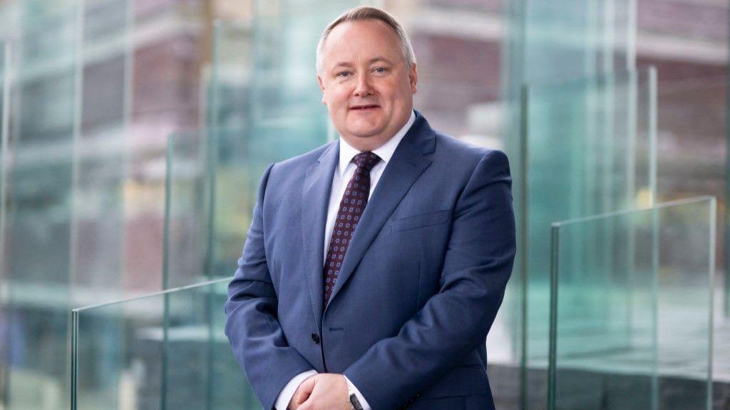 A picture of Darren Millar on the Senedd steps, standing in front of a glass sculpture. He is wearing a blue suit, a red spotted tie and a white shirt.