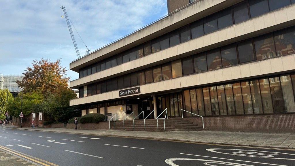 Outside a courthouse. There is a set of steps, white handrails, and glass windows outside, and a black sign with silver lettering saying "Seax House" above the entrance.