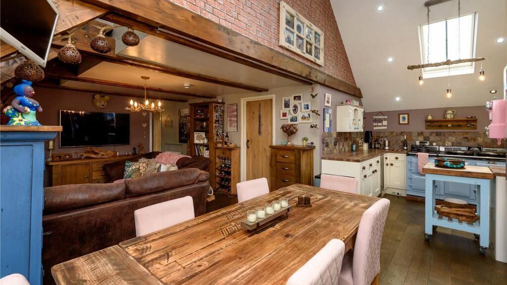 The kitchen and a living area of the property which accompanies the Polperro Model Village, which is open plan, some some exposed brickwork, a large wooden dining table and country-style kitchen