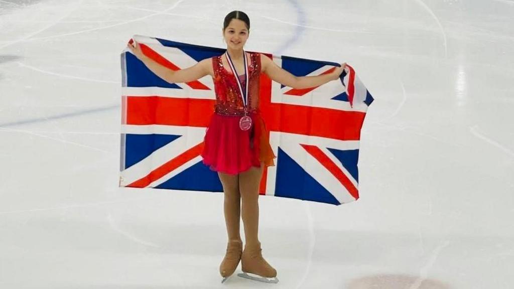 Leilah is on the ice with a British flag held behind her back. She is smiling. 