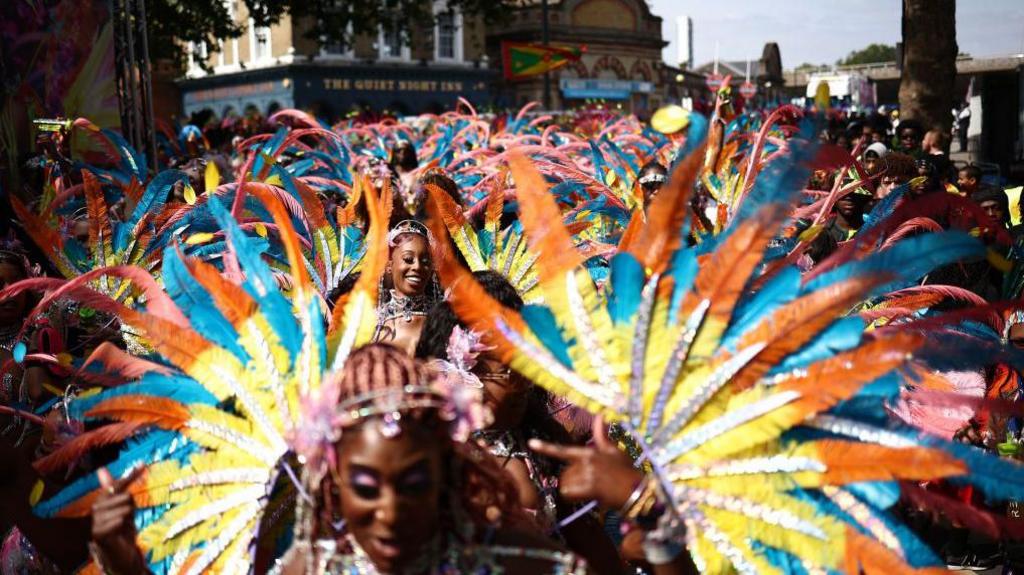 Performers wearing elaborate feathered headdresses in bright orange, blue and yellow take part in a Notting Hill Carnival parade, with crowds of spectators lining the street in the background.