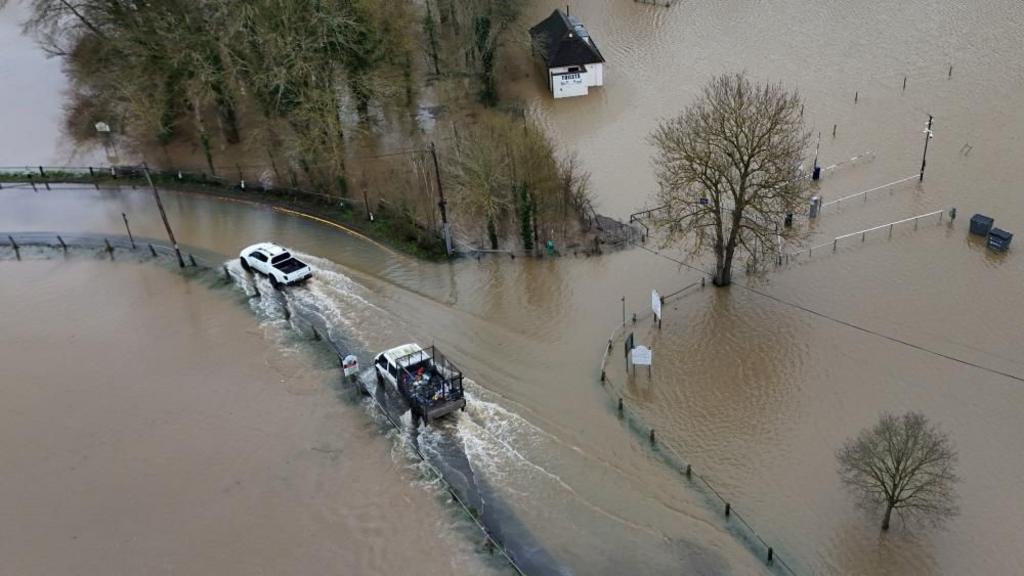 From above, two cars drive through brown floodwaters as a small house can be seen surrounded by water