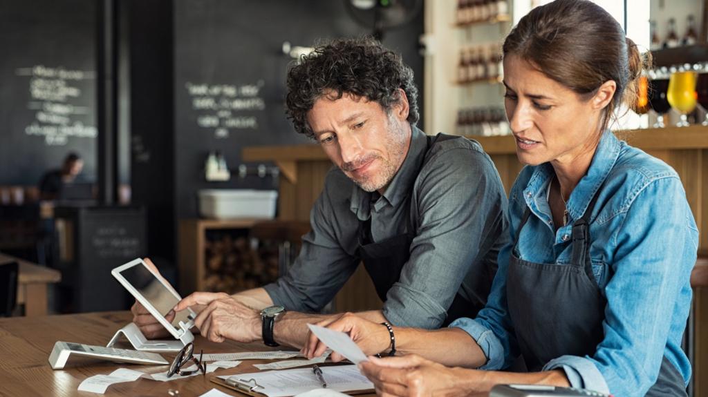 A man and woman - both wearing smart shirts and black aprons - sit at a restaurant table looking at bills. The man has his finger scrolling through a tablet.