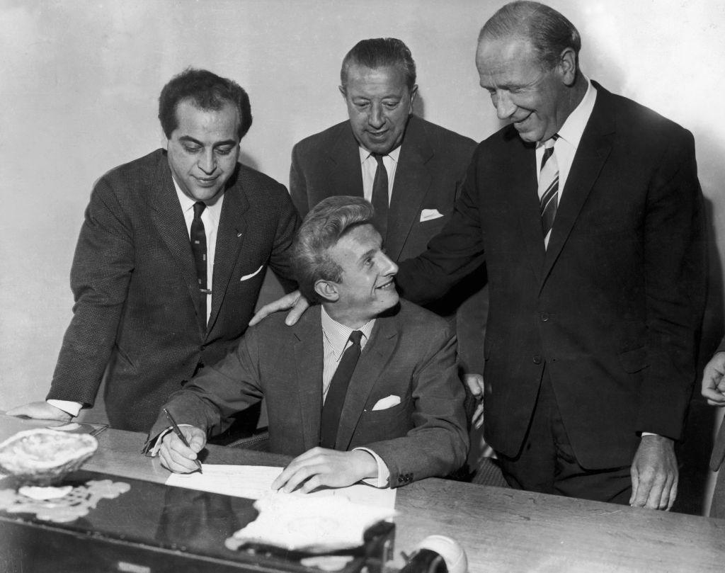 Four men wearing dark suits in a black and white picture. One man, Denis Law, sits at a table signing a contract while the others gather behind him. Everyone is smiling.