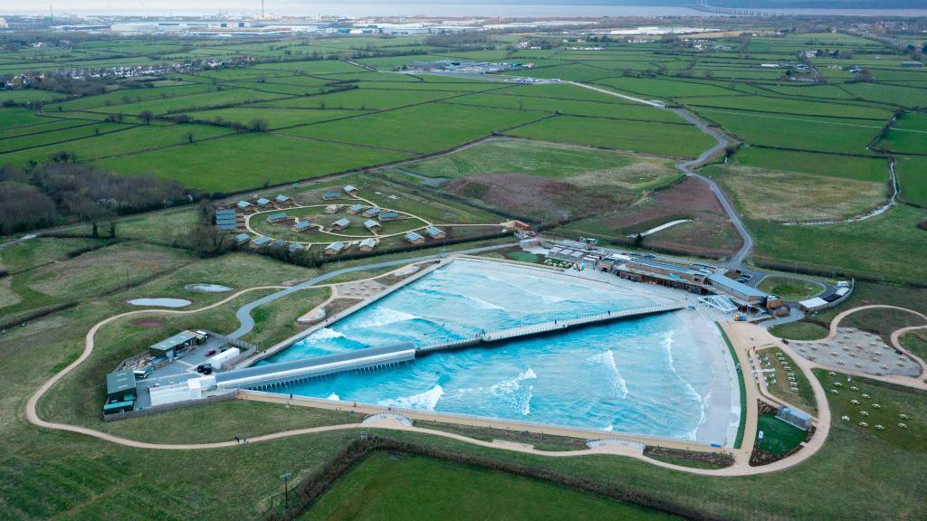 An aerial view of an artificial wave pool in a rural setting, surrounded by green fields, pathways, and small buildings, with an industrial area and a bridge in the background.