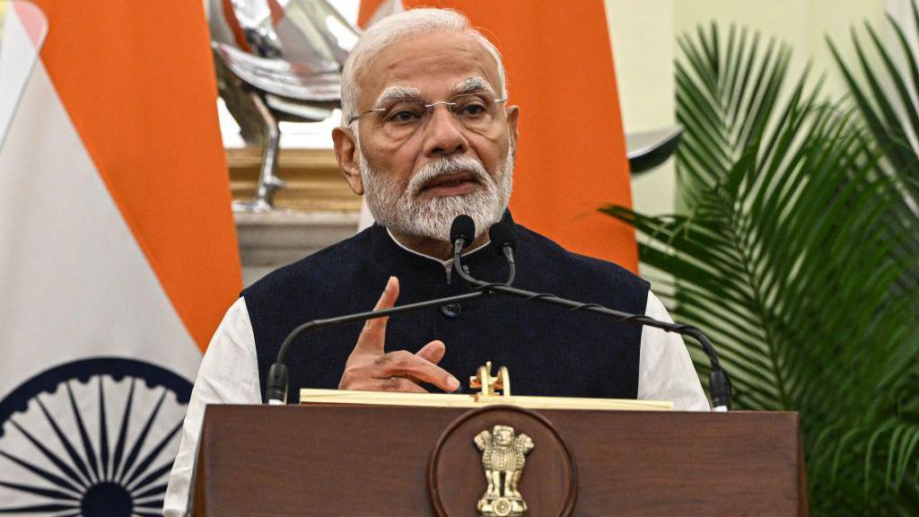 Narendra Modi, wearing a white long-sleeved shirt and black vest, stands before a microphone at a wooden lectern with a gold seal, in front of an Indian flag, and holds his finger up as he speaks