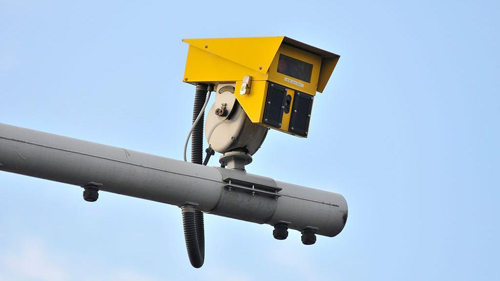A close up of a yellow average speed camera resting on a grey pole, against a blue sky