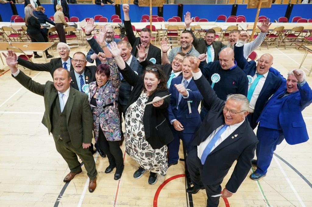 Reform candidates celebrate the Hartlepool election result. There are a group of 16 men and woman standing close together in a group with their hands raised in the air and celebrating. Many of them are wearing light blue and white rosettes. They are standing in a leisure centre with markings on the floor and a row of desks and chairs behind them