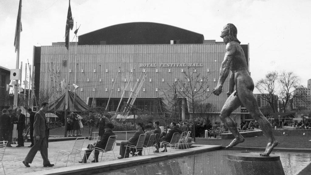 Black and white image of a male nude statue stand by a pool of water in front of the newly constructed Festival Hall in 1951. Some people are sitting in chairs in front of the pool. 