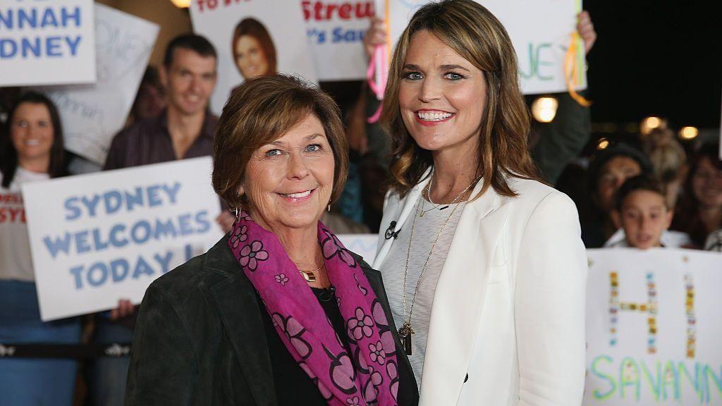Savannah, all in white, and Nancy Guthrie in a black suit jacket and pink patterned scarf, smile as they stand shoulder to shoulder. People behind them hold up signs. 