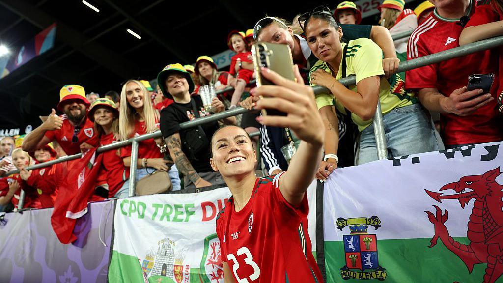 Ffion Morgan in a Wales shirt takes a selfie with Wales fans behind her higher up in the stand. Wales flags are draped over the barrier.