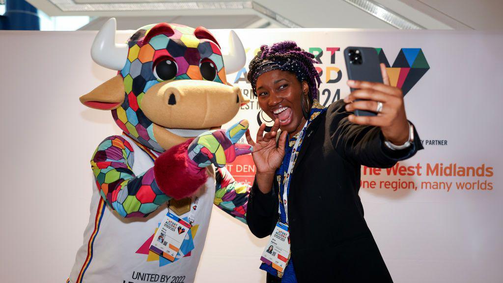 a multicoloured bull mascot taking a selfie with a woman at a convention