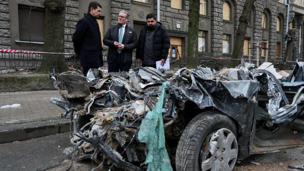 The prime minister guided by Ukrainian officials as he inspects a damaged vehicle along a street