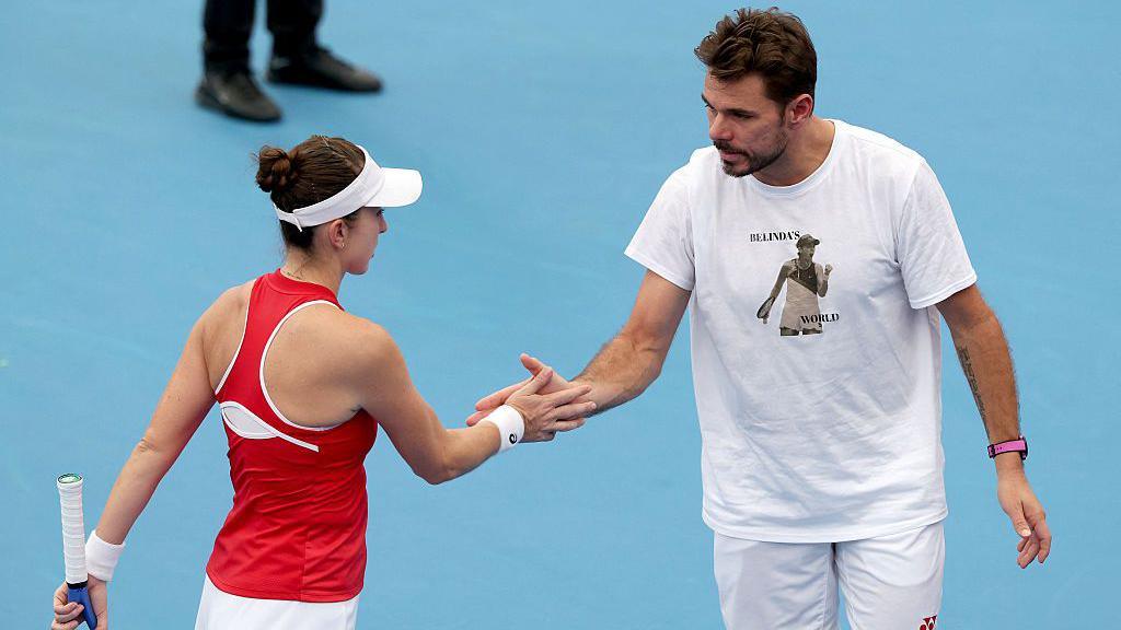 Switzerland's Belinda Bencic, in a red tennis top and white visor, reacts with team-mate Stan Wawrinka as they touch palms. Wawrinka is wearing a white T-shirt with a picture of Bencic and the words BELINDA'S WORLD