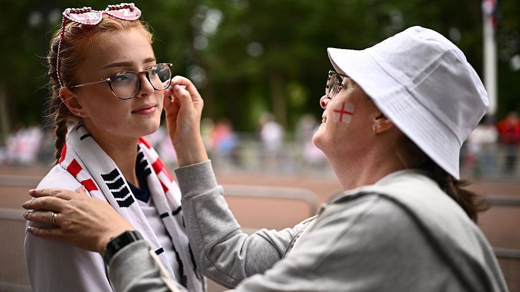 Girl wearing glasses is seen having faceprint applied to her cheek by a woman who is wearing a white hat.