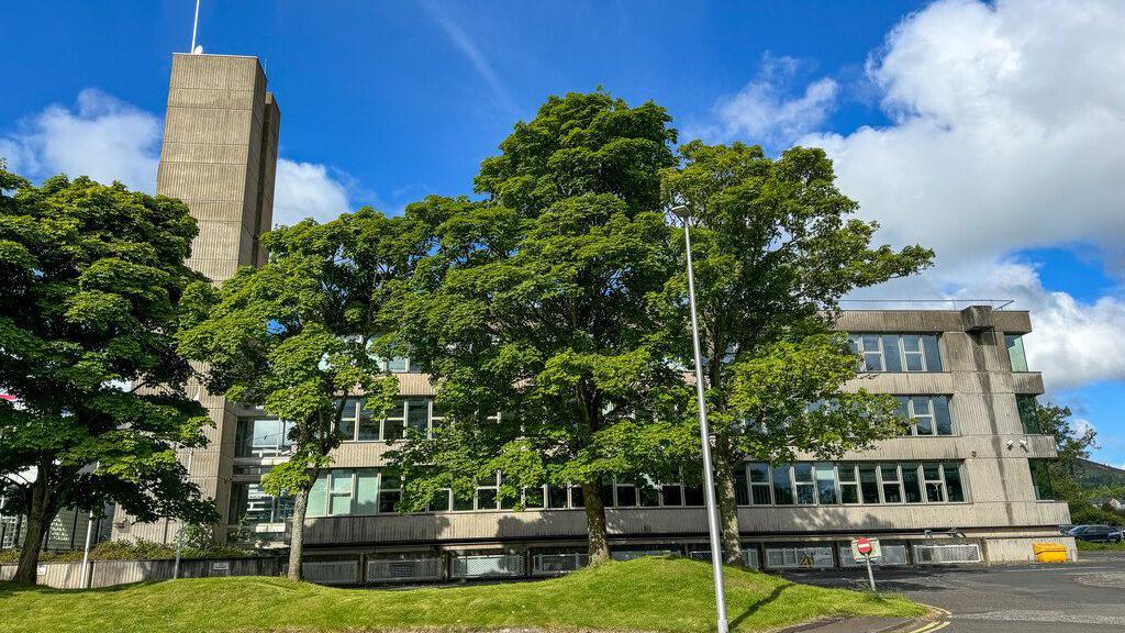 A grey concrete building with a tower concealed in large part by a large tree