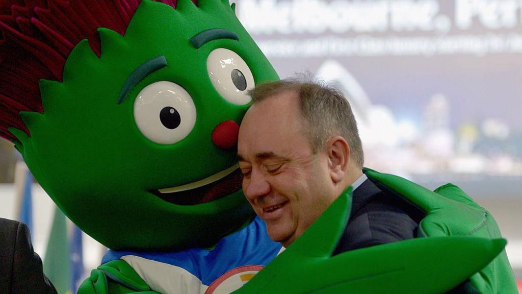 Clyde the green and purple animated thistle hugging Alex Salmond, who has his eyes closed and is smiling
