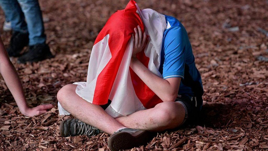 An England fan sits on the ground with holding his head with his hands. He is hiding his face with an England flag over his head.