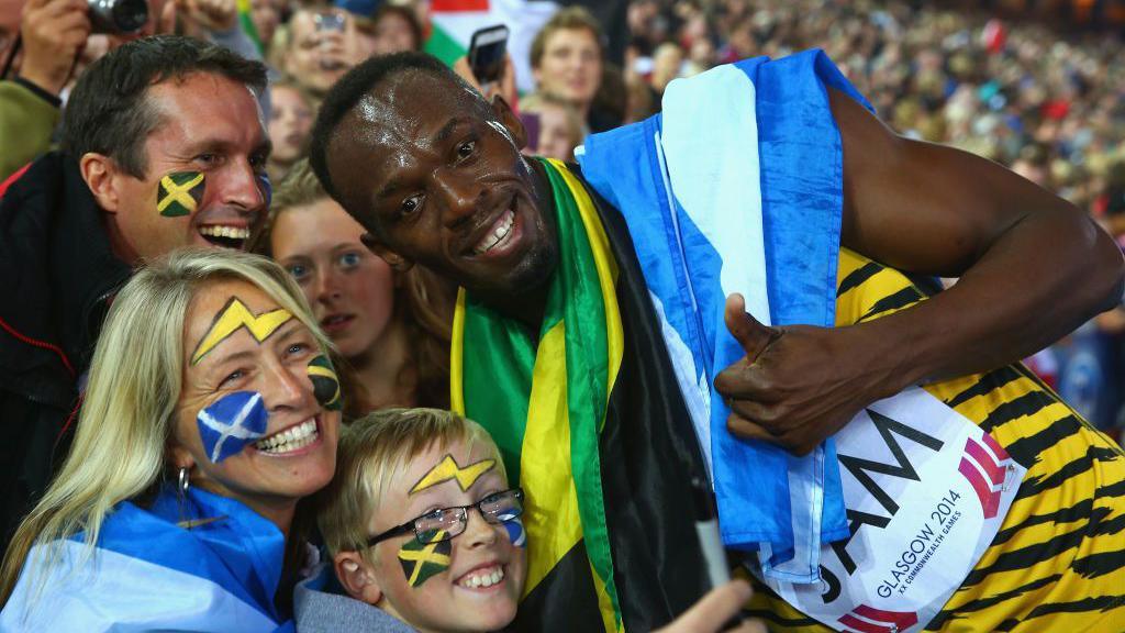 Usain Bolt celebrates with fans after winning a medal at the Commonwealth Games. Bolt is wearing his running gear, and draped with the flags of Jamaica and Scotland. He is grinning and giving a thumbs up as he poses with a mum and a young boy - she has a Scotland flag wrapped around her and both fans have their faces painted with the Jamaican and Scottish flags, plus lightning bolts.