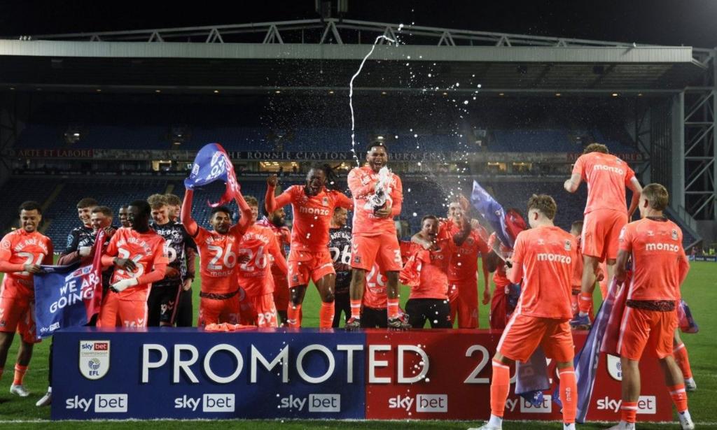 Coventry City are lined up on the pitch at Ewood Park standing behind a display banner at their feet that says Promoted 2025/26. The players are euphoric in victory, with spray from a champagne bottle shooting up above them.