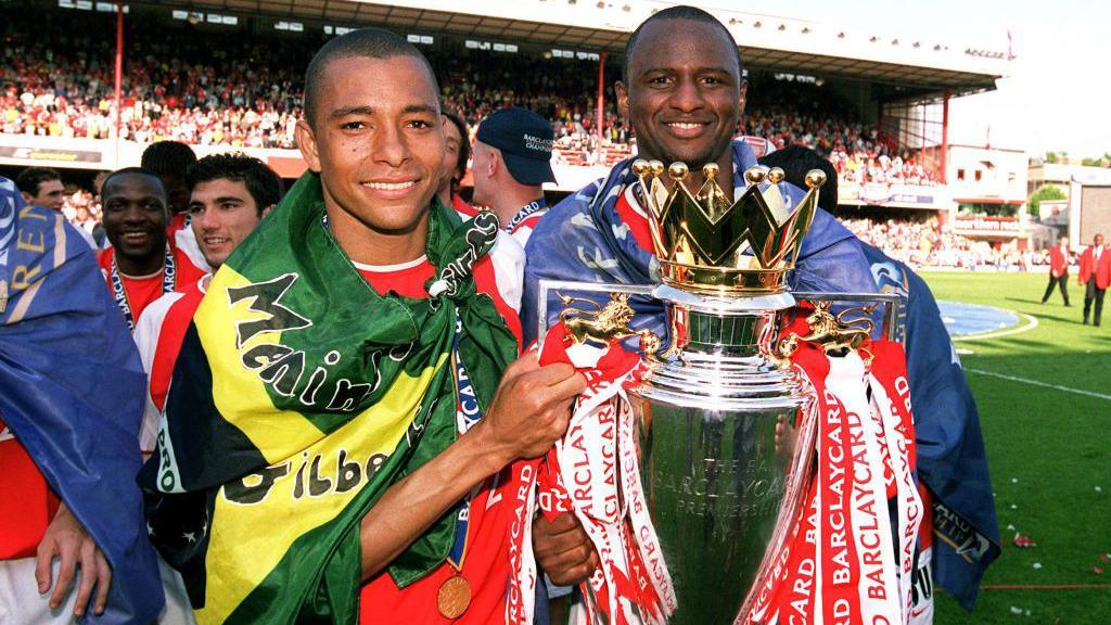 Gilberto Silva [left] wears a Brazil flag as he lifts the Premier League trophy with Arsenal captain Patrick Vieira 