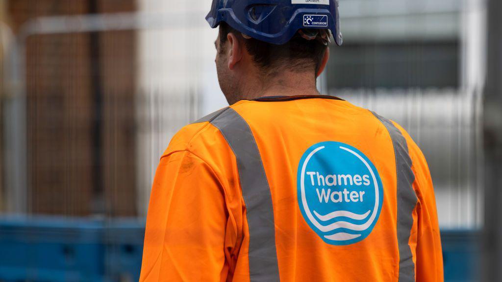 Thames Water worker wearing a navy hard-hat and an orange high vis jacket featuring the company logo