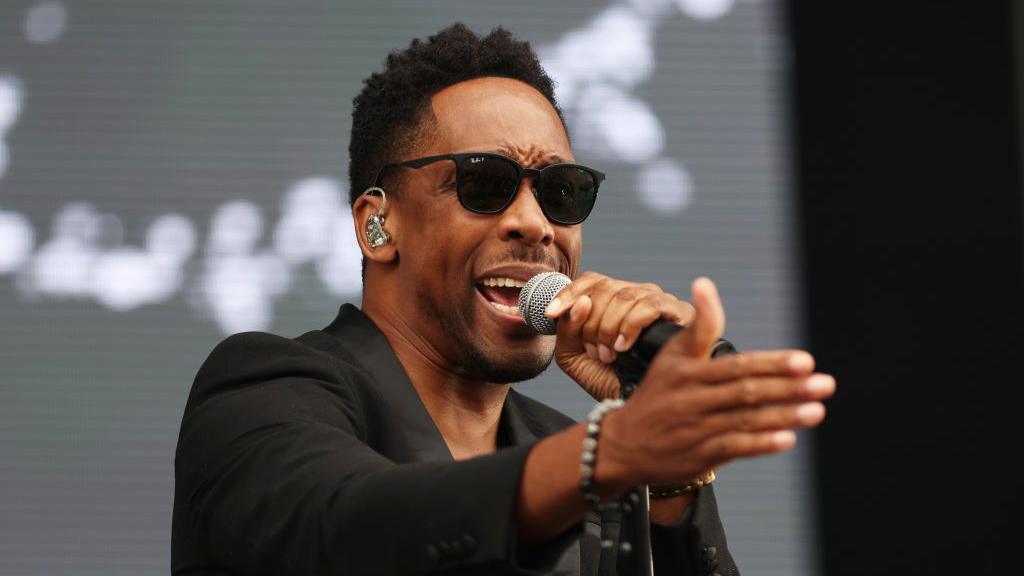 Lemar performs at BBC Radio 2 In The Park 2023 at Victoria Park in Leicester, England. He wears a black tuxedo jacket, black sunglasses and sings into a microphone. He has cropped afro hair and wears a silver bracelet.