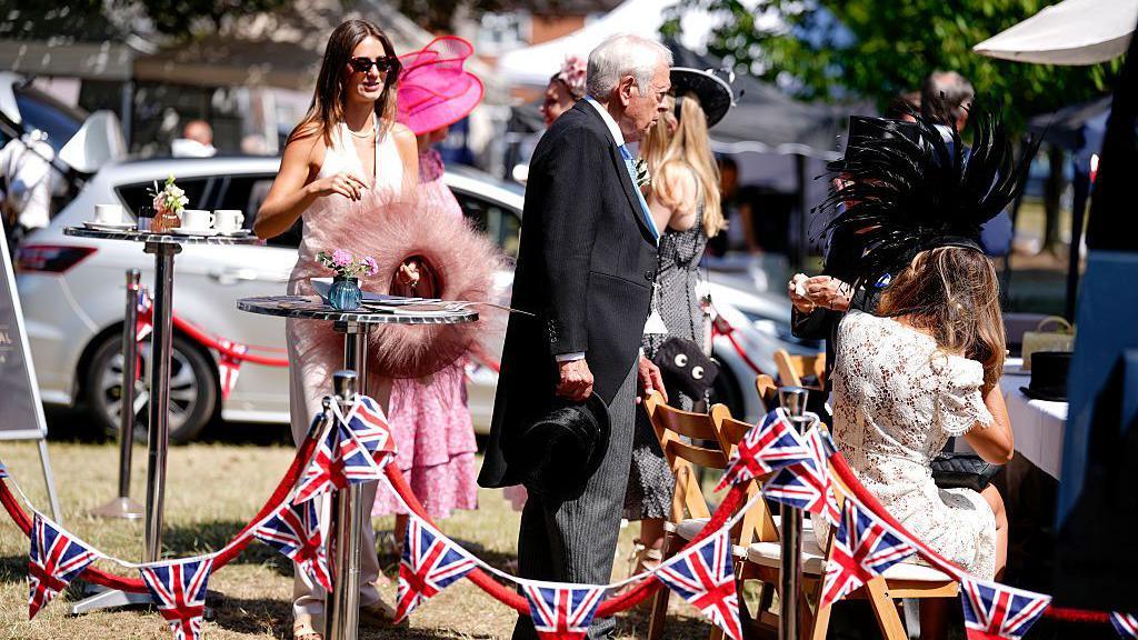 A man in a tailcoat suit and a group of women dressed in formal dresses and hats standing or sitting around a table in the sun, with Union Jack bunting next to them.