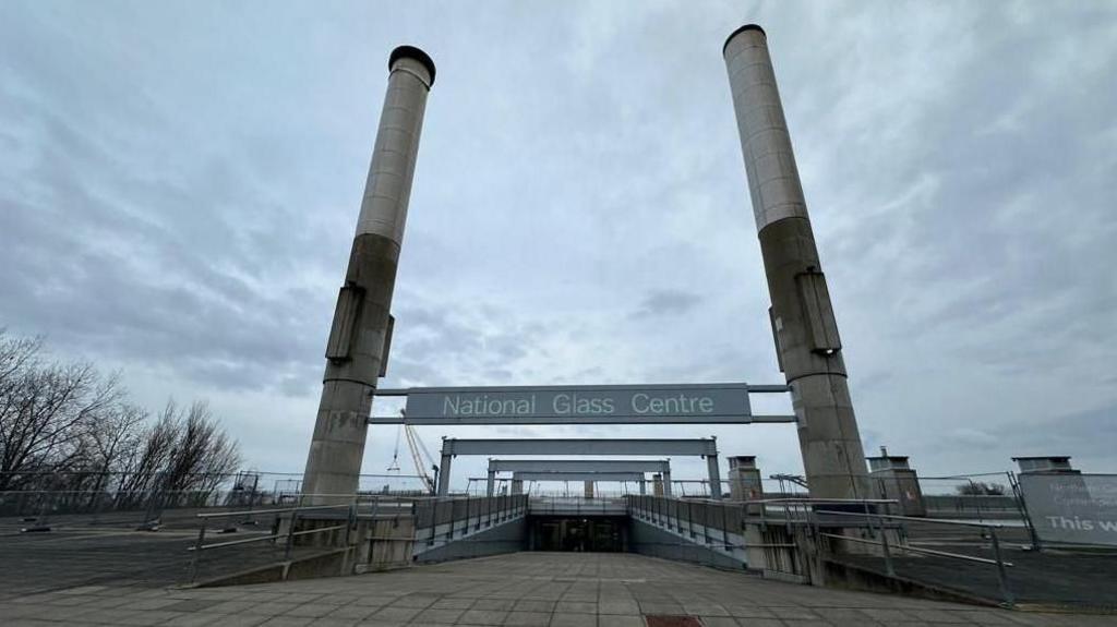 The entrance to the National Glass Centre. The concrete path leads down a ramp. Meanwhile, two concrete pillars border either side. A grey sign reads: "National Glass Centre".
