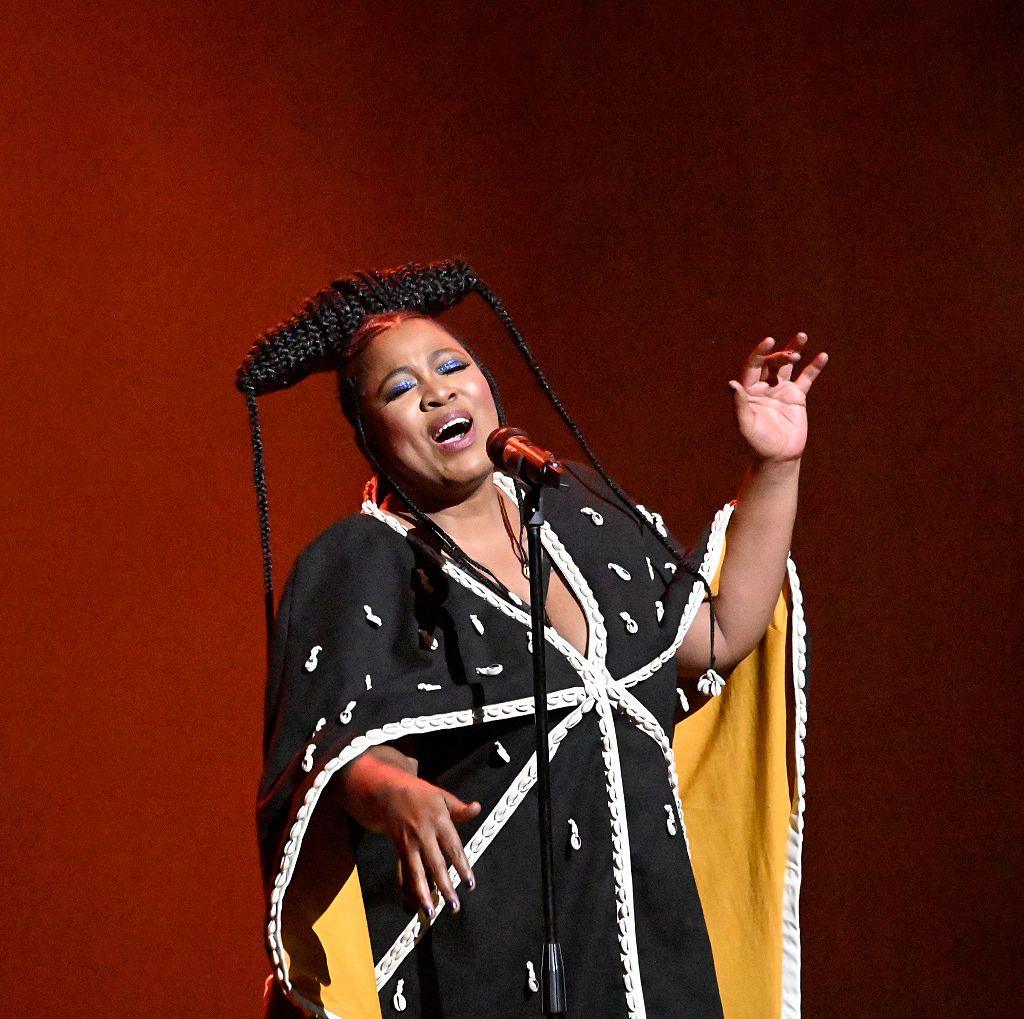 A woman sings on stage wearing a black and white robe which is beaded with cowrie shells.
