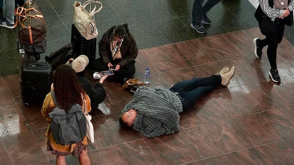 man lying on floor wrapped in blanket. near him, sits a woman cross legged on her phone and a suitcases next to her. There is another woman wearing a backpack with her back to the camera