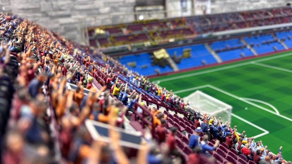 Tiny plastic figures sat on plastic chairs in a model of Villa Park. The green pitch can be seen on the right with a football net, and more seats in the background.