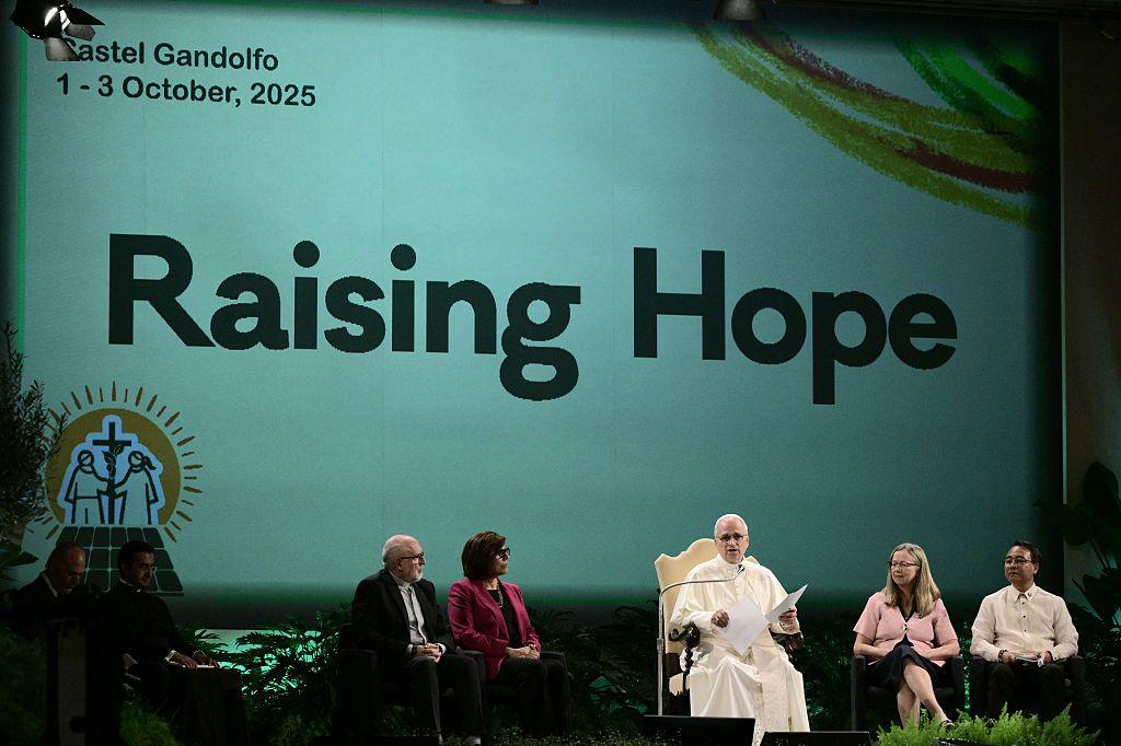 On a stage in his summer residence, Castel Gandolfo, the Pope dressed in white, speaks to an audience. Four other speakers sit beside him, listening intently. 
