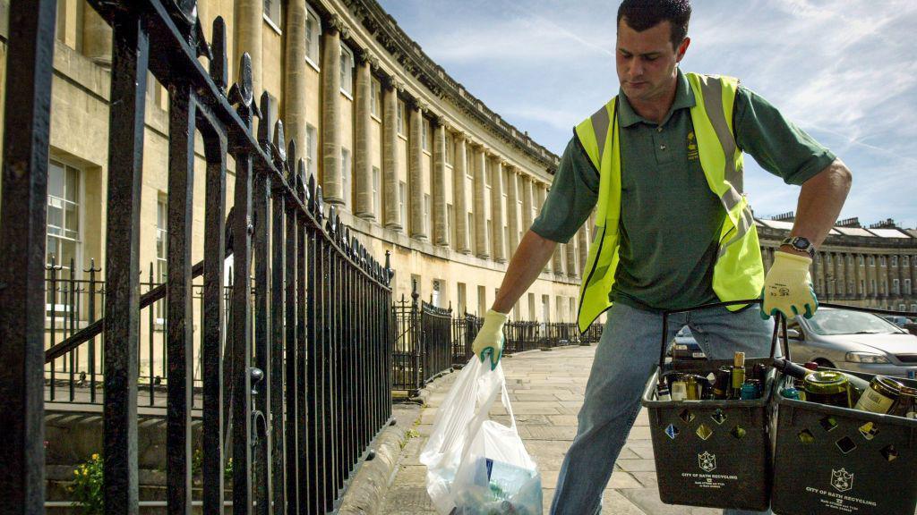 A man with brown hair, wearing a yellow and grey high-visibility vest, gloves and a green shirt stands on Bath Crescent holding two black City of Bath recycling containers filled with glass bottles and a plastic bag of general waste, with terraced buildings visible behind him.