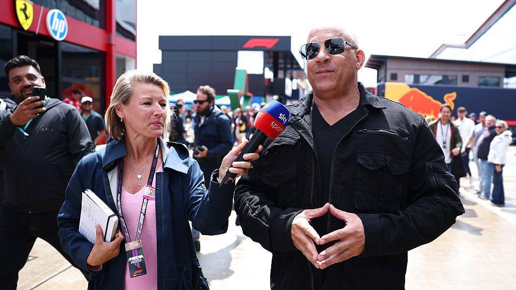 Vin Diesel being interviewed. He is wearing a black jacket and black top and black sunglass with a smile on his face. The female interviewer is wearing a pink top and blue coat. An onlooker takes a picture on their mobile phone. They are all in a busy paddock area.